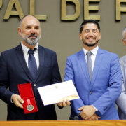 O presidente da CEASA/RS, Carlos Siegle, recebe a Comenda Porto do Sol no plen&aacute;rio da C&acirc;mara Municipal de Porto Alegre. Ao centro da imagem, ele segura a medalha da honraria e o certificado ao lado do vereador Giovane Byl, autor da homenagem, enquanto autoridades acompanham o momento durante a solenidade.